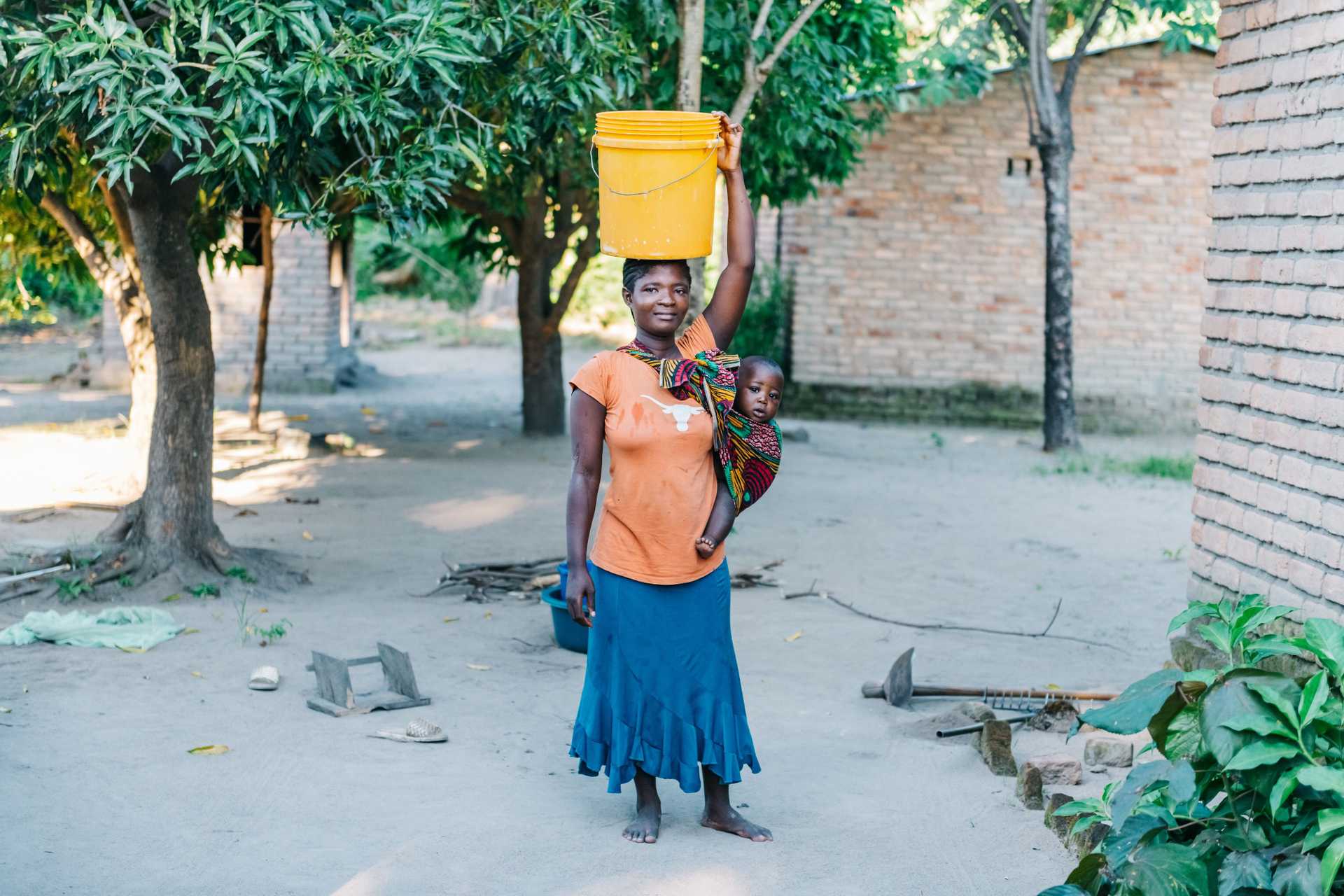 young african mother with baby carrying water bucket on head in village in Malawi